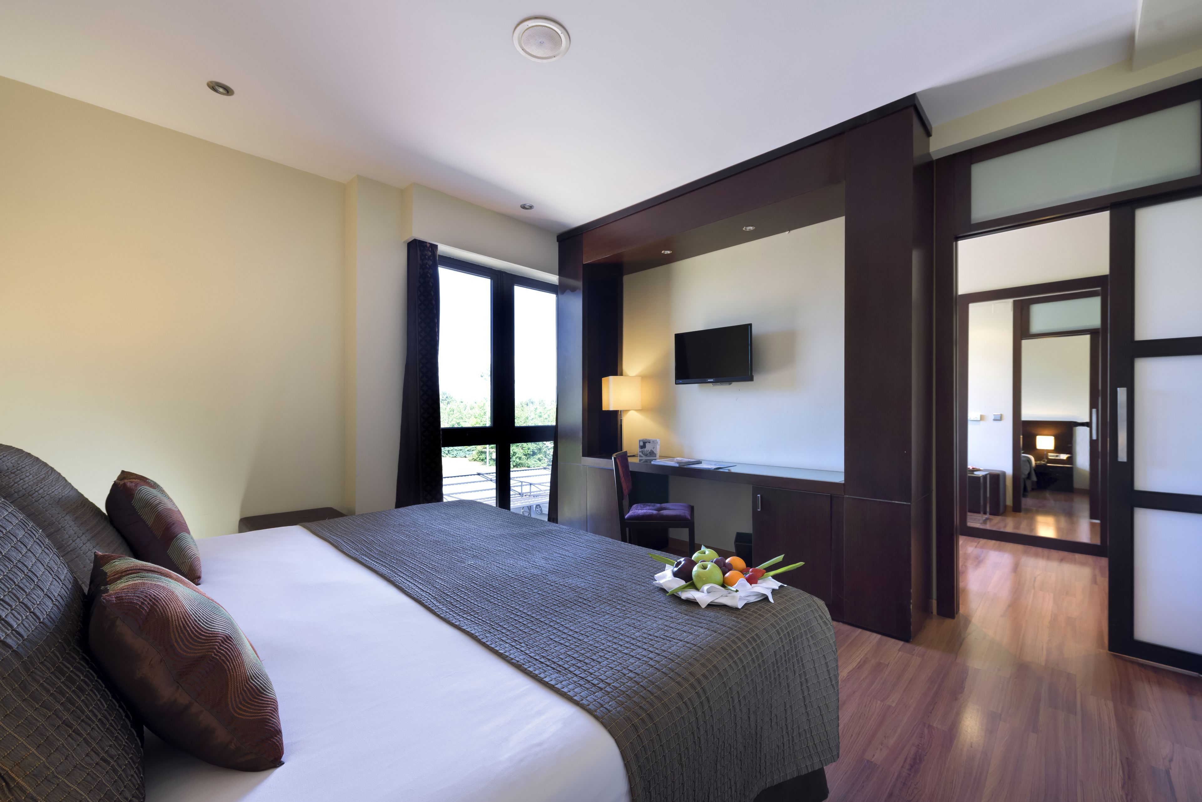 Ein Hotelzimmer mit einem großen Bett, einem Schreibtisch mit Stuhl, einem an der Wand befestigten Fernseher und Blick ins Freie durch ein Fenster.