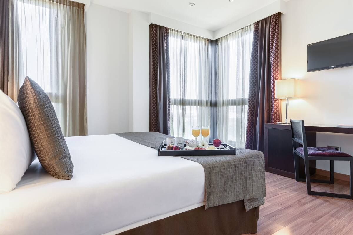 A hotel room with a bed, a tray with breakfast items, a desk with a chair, and a TV mounted on the wall.