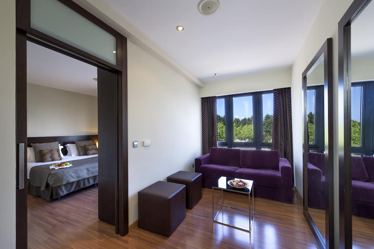 A modern hotel room with a large bed, a purple sofa, a small table, and a large window offering a view of the outdoors.