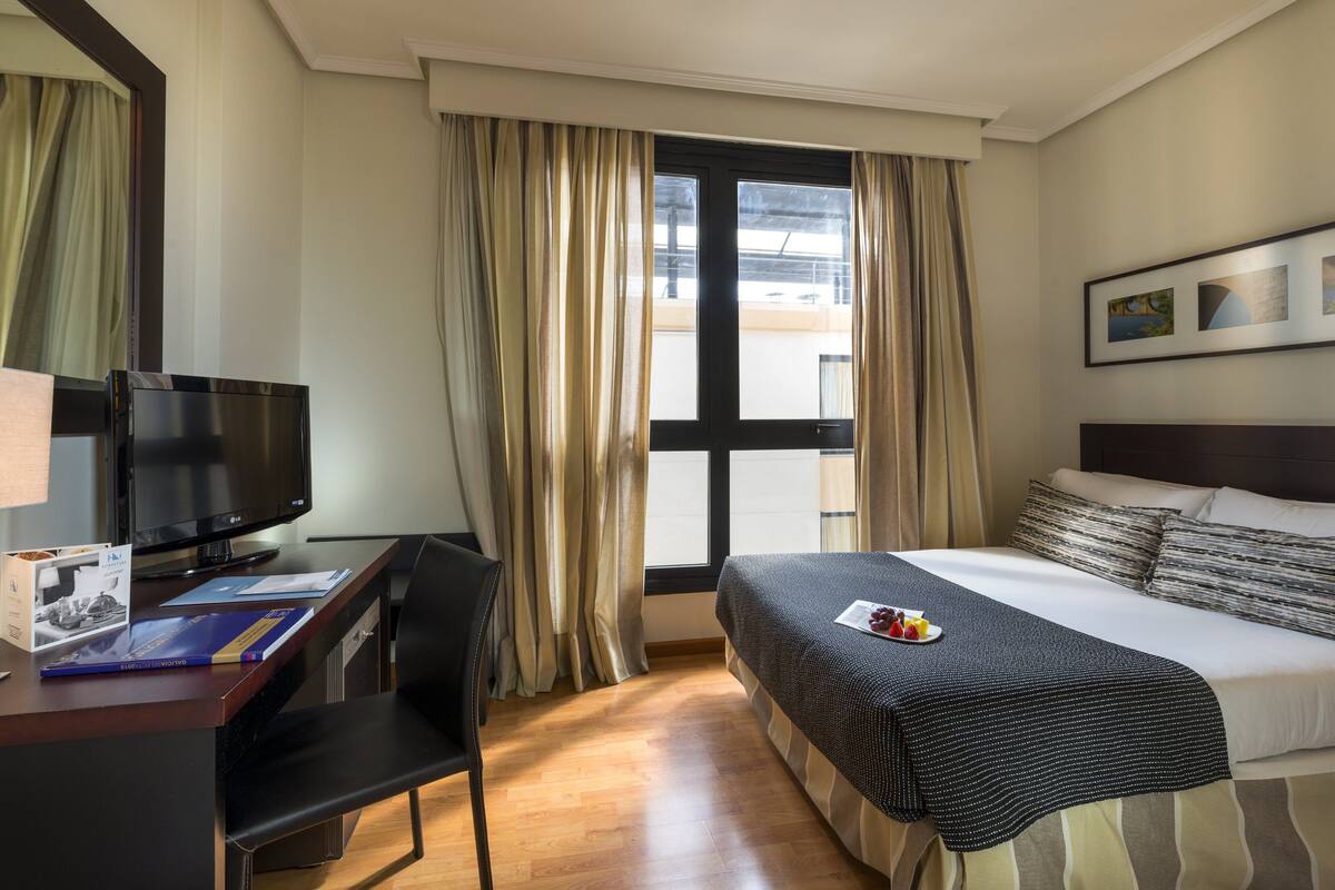 A hotel room with a bed, a desk with a computer, a chair, and a window with curtains.