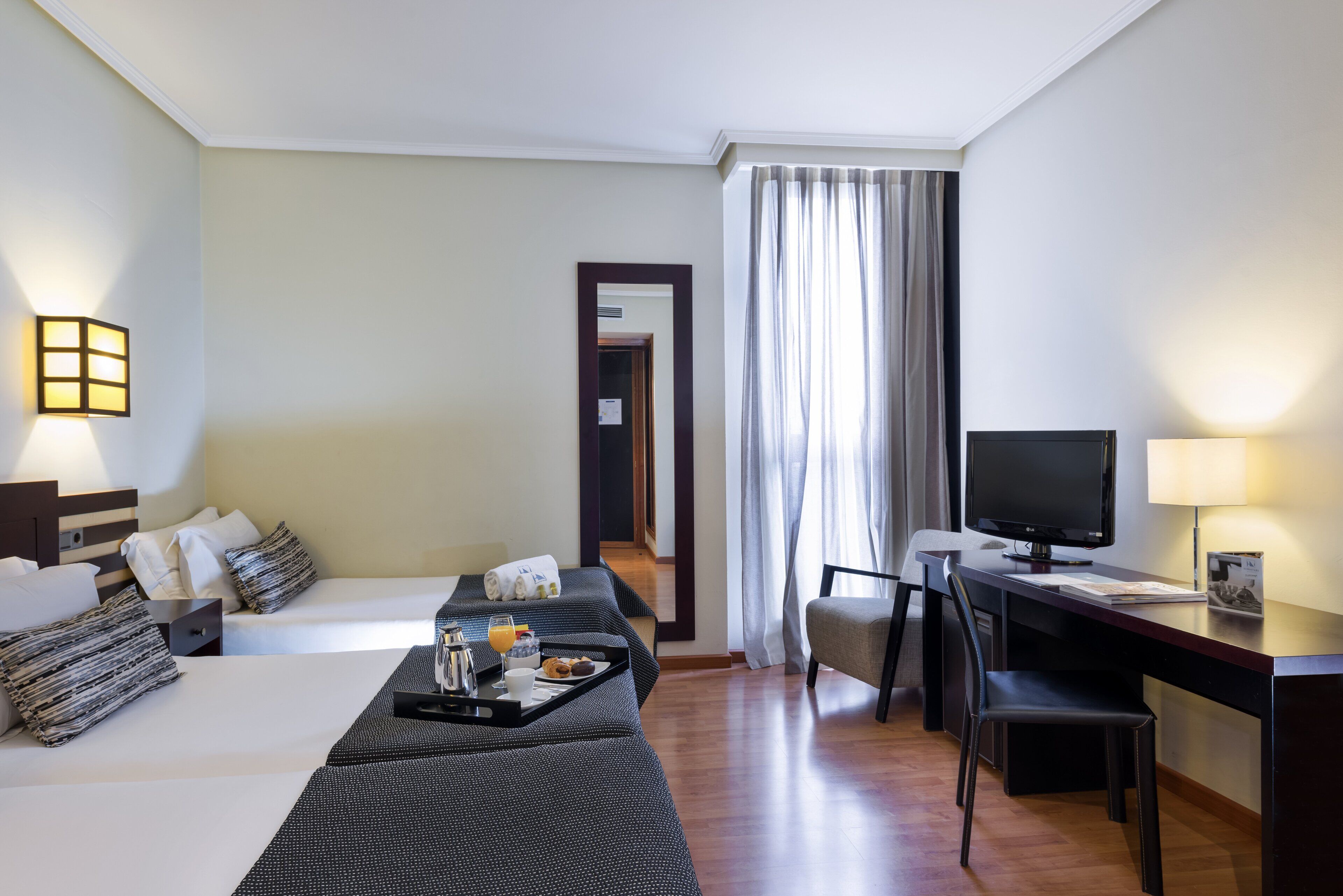 Une chambre d’hôtel avec deux lits, un bureau avec un ordinateur, une chaise, une télévision et un plateau avec des articles pour le petit-déjeuner.