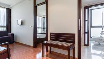 Ein modernes Hotelzimmer mit Holzboden, einem dunkelgrauen Sofa, einer Holzbank, einem großen Spiegel und einem Badezimmer mit weißem Waschbecken und Badewanne.