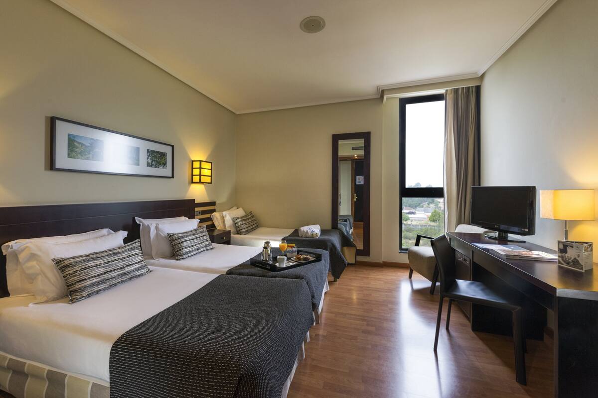A hotel room with two beds, a desk, a chair, a TV, and a window with a view.