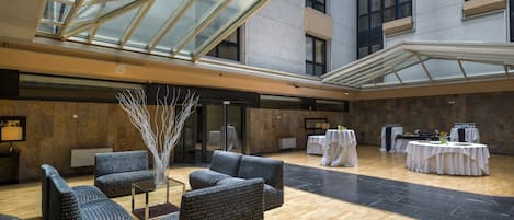 Ein großzügiger Innenbereich mit hoher Decke und Glasdach, ausgestattet mit einer Sitzecke mit Sofas und einem zentralen Dekorationsstück. Im Hintergrund sind Esstische aufgestellt.