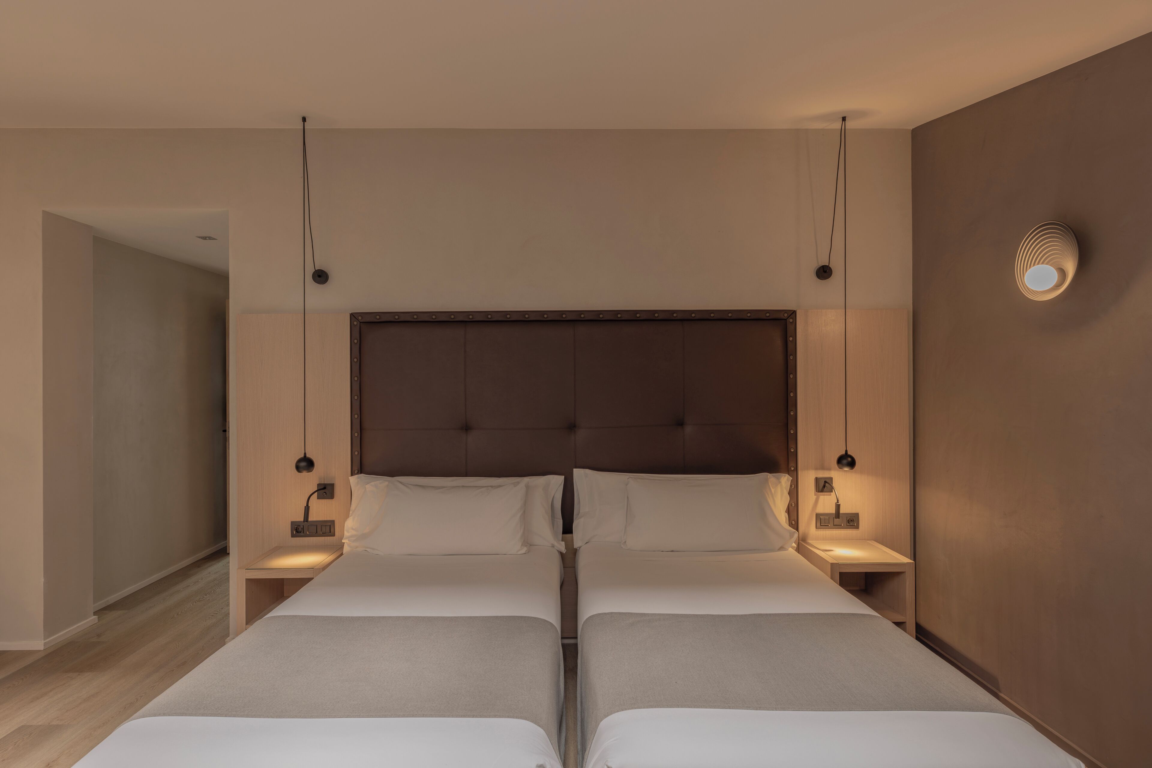 Habitación de hotel con dos camas, cabecero de madera, lámparas de noche y apliques en la pared.