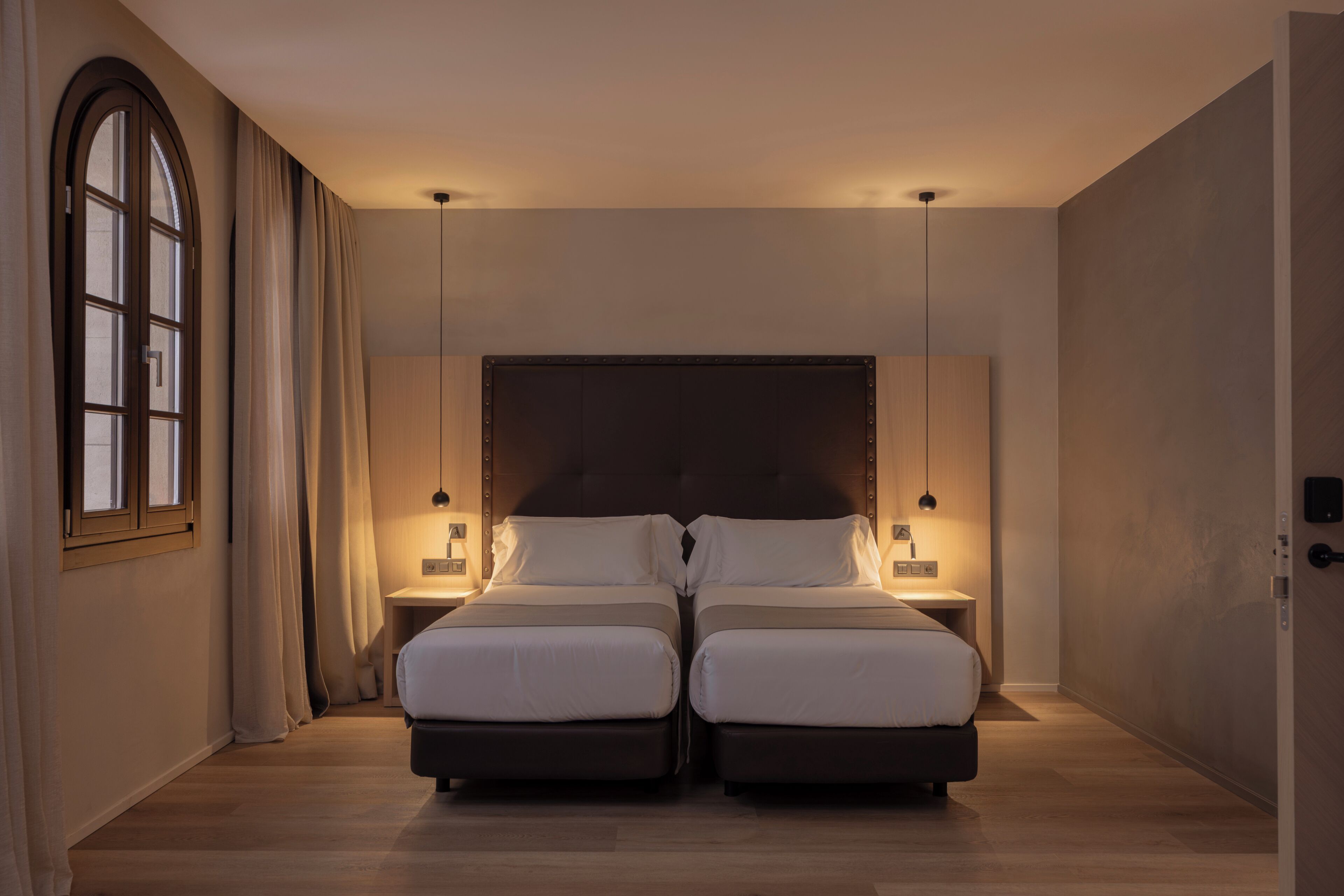A double bed with a dark headboard, two bedside tables with lamps, a large arched window, and a wooden floor.