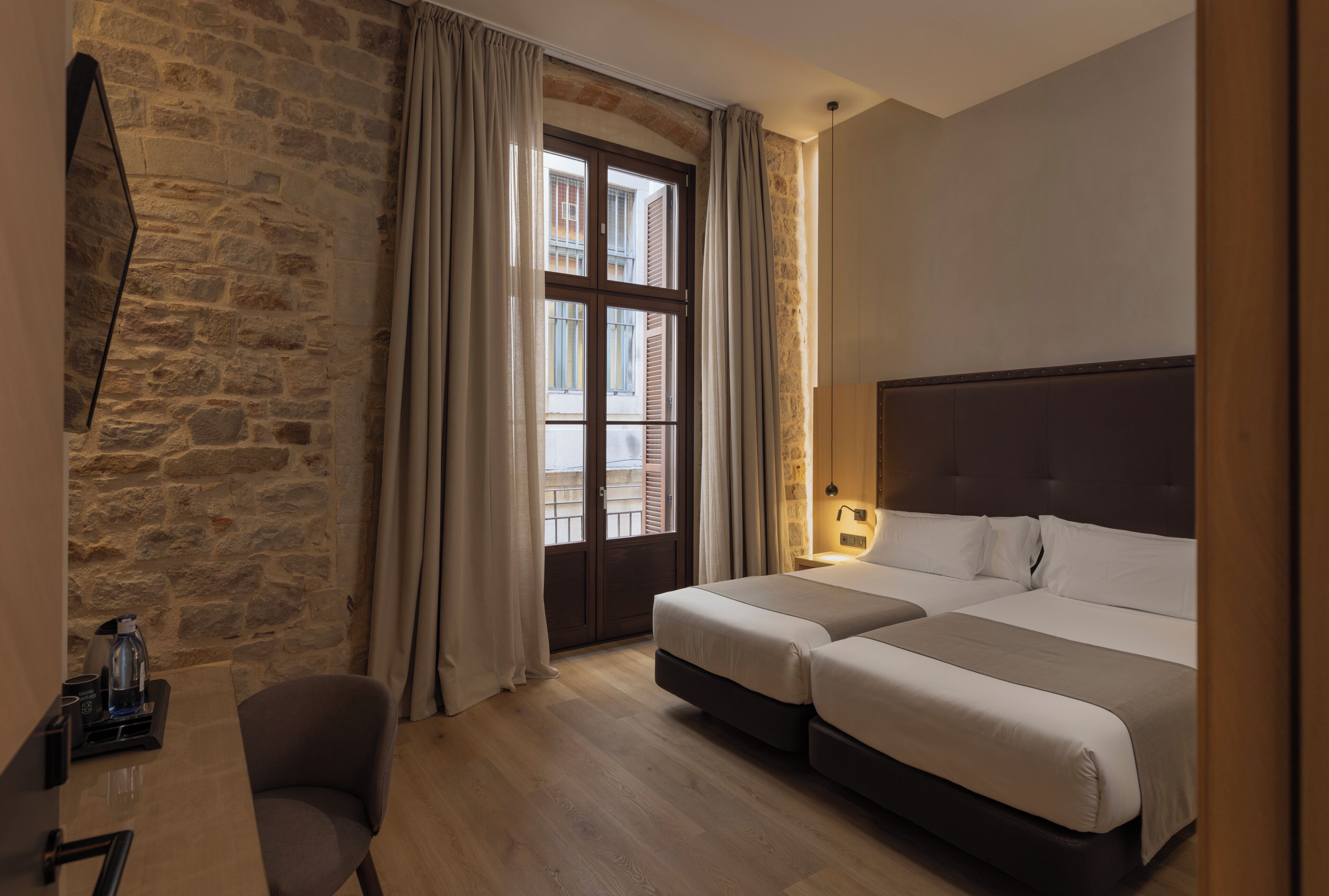 A hotel room with a stone wall, a bed with two pillows, a chair, a desk with a coffee maker, and a window with curtains.