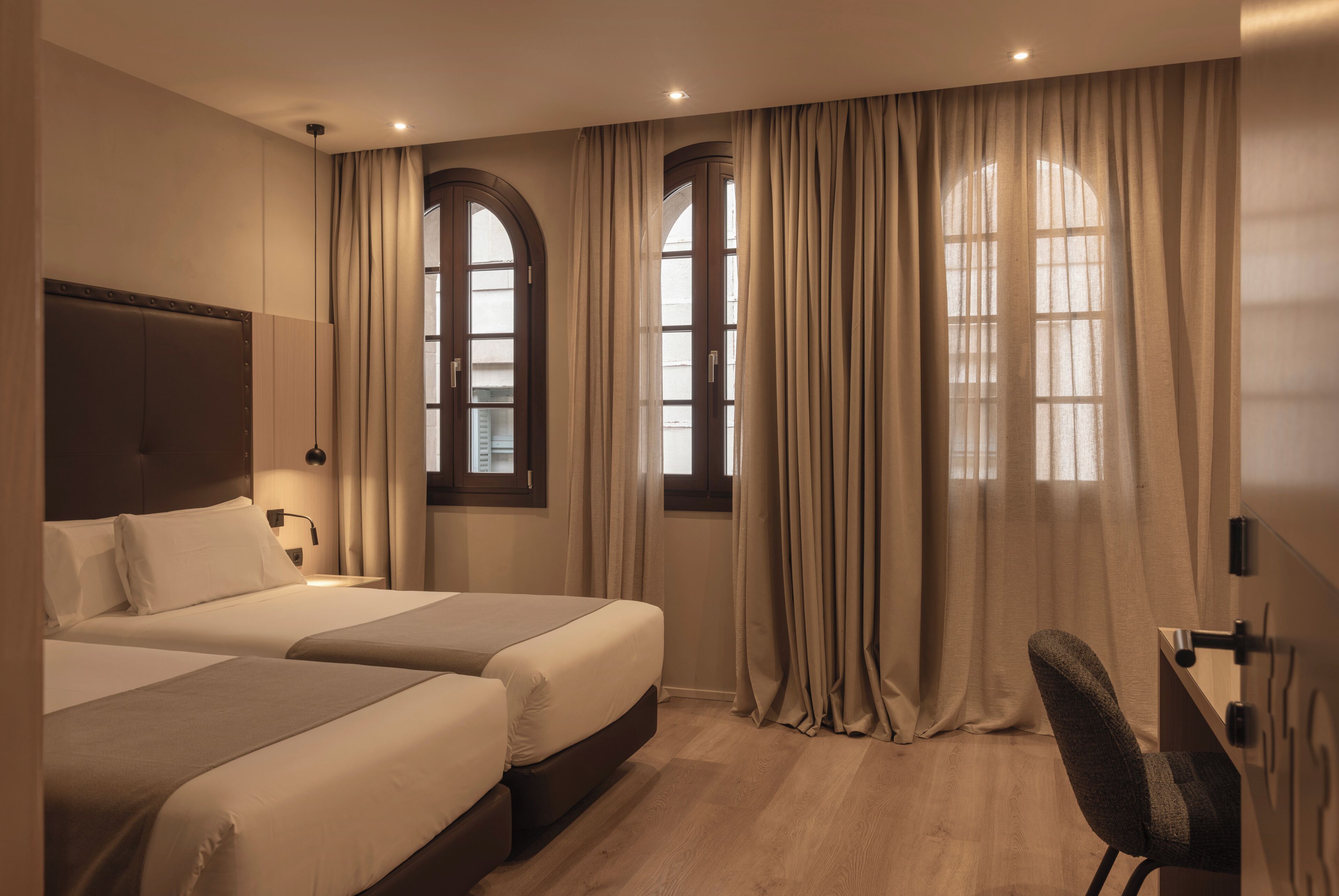 A hotel room with two beds, a desk, and large windows with curtains.