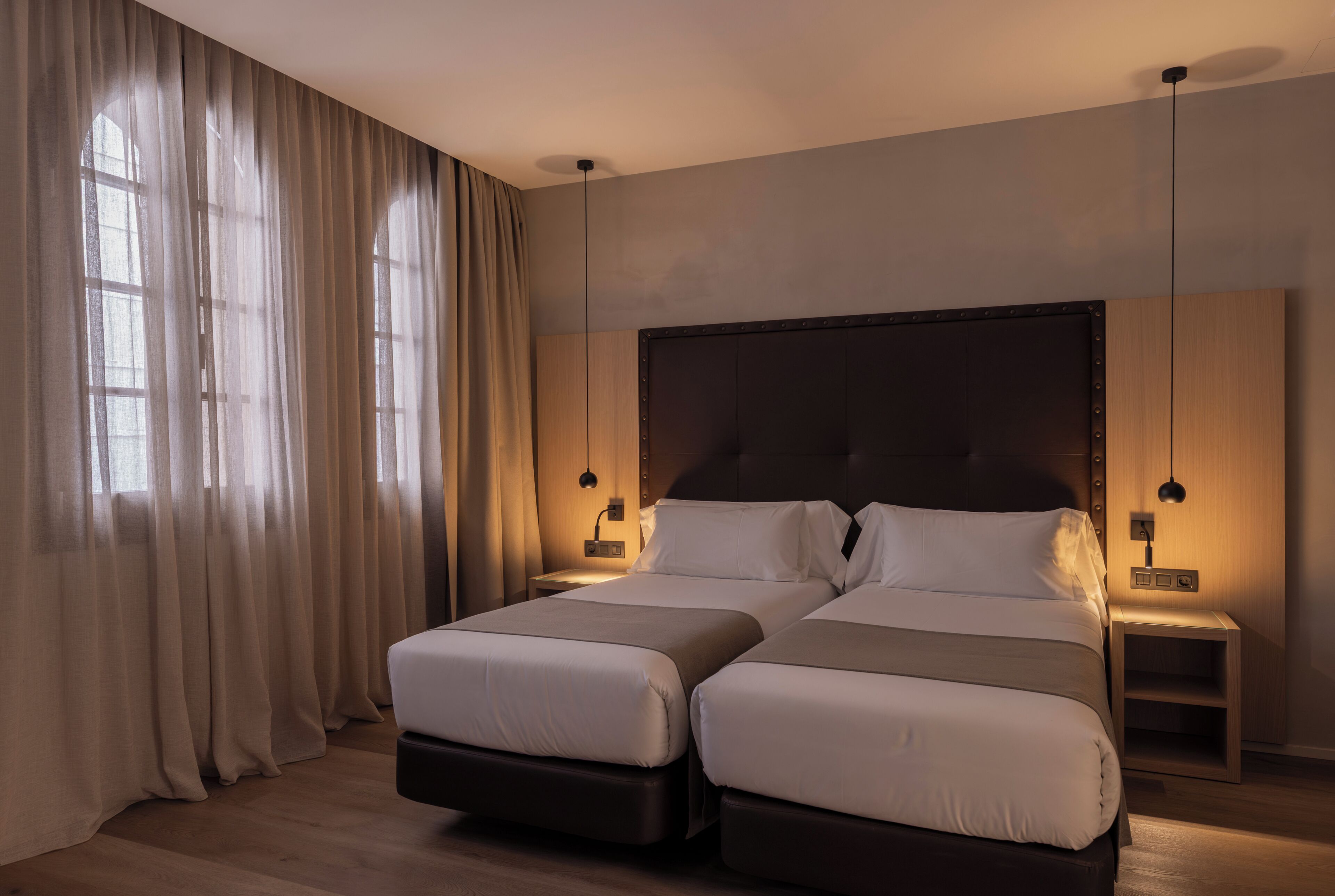 A double bed with a dark headboard, two bedside tables, and a large window with sheer curtains.
