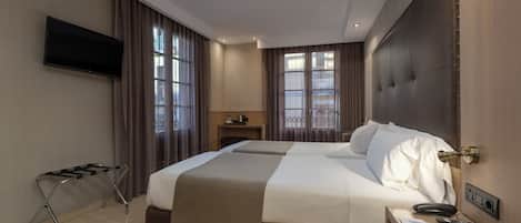A hotel room with a large bed, a TV mounted on the wall, a bedside table with a phone and a lamp, and a window with curtains.