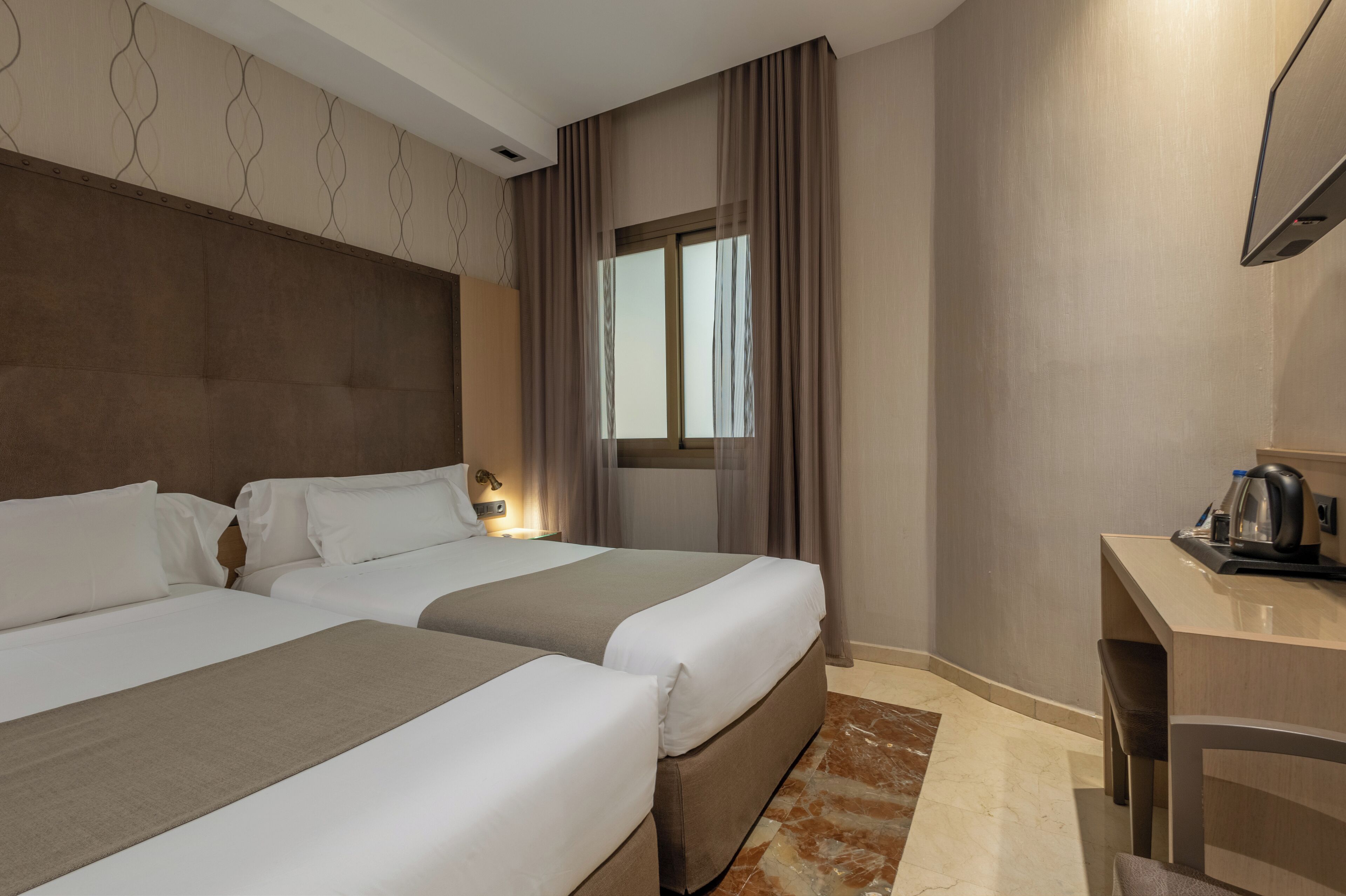 Habitación de hotel con dos camas, un escritorio, una silla, una ventana con cortinas y un televisor montado en la pared.