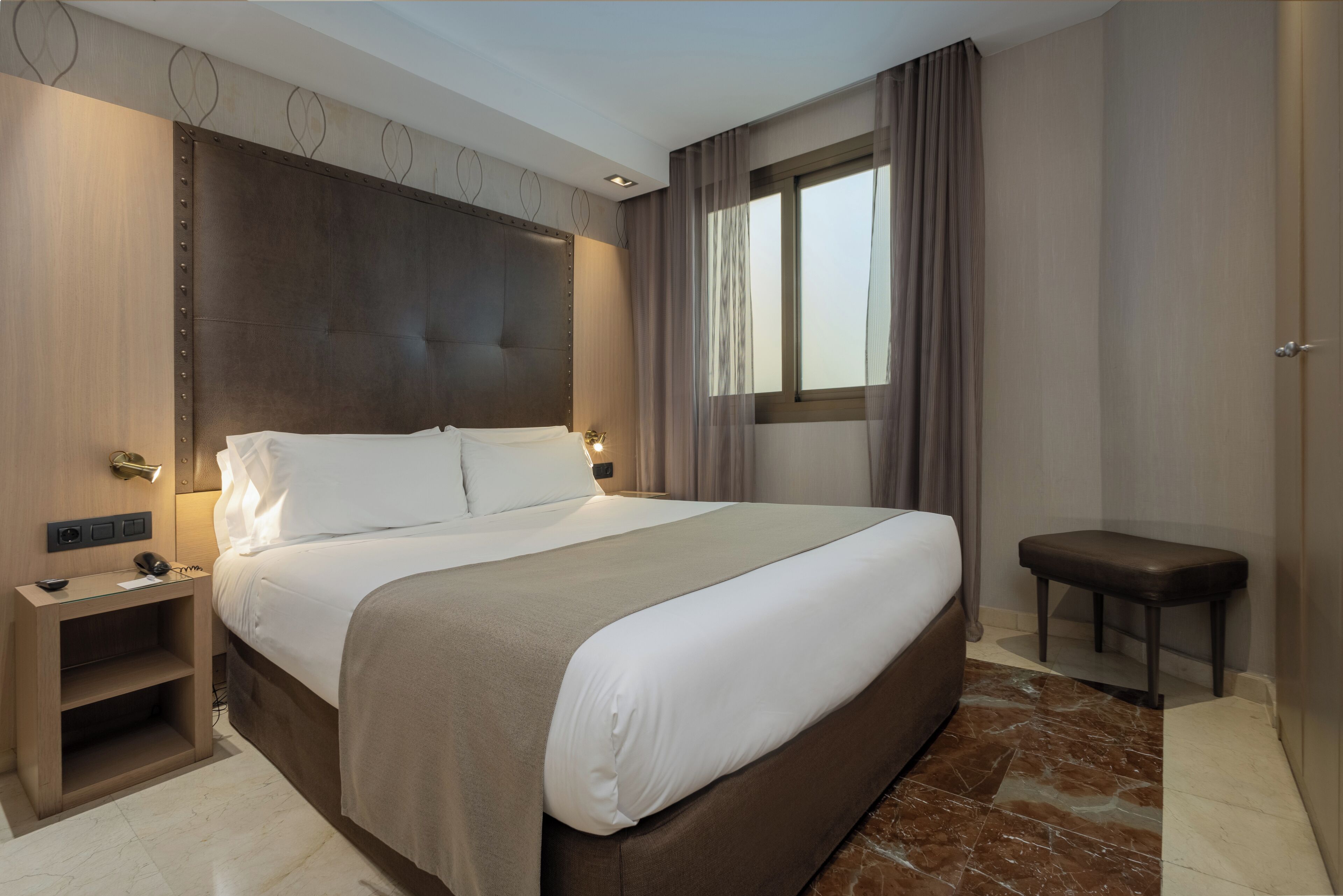 Une chambre d’hôtel moderne avec un grand lit, des tables de chevet, un petit banc et une fenêtre avec des rideaux.