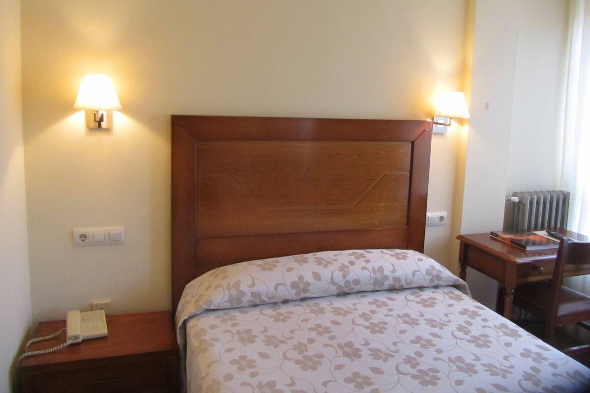 A hotel room with a wooden headboard, a bed with floral bedding, bedside lamps, a telephone, and a small table with a book and a lamp.