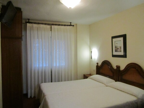 A bedroom with a bed, curtains, a nightstand, a wall-mounted light, and a framed picture.