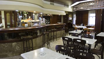 A restaurant interior with marble tables, dark wooden chairs, a bar area, and a high ceiling with decorative moldings.