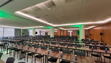 Een ruime congreszaal met rijen tafels en stoelen, voorzien van waterflesjes, en een podium met een spreker en scherm.