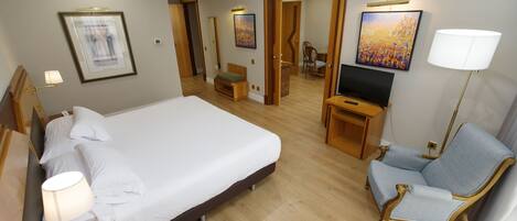 A hotel room with a large bed, a TV, a chair, and artwork on the walls.