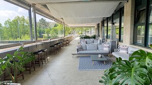 A covered outdoor seating area with a sofa, chairs, and tables, surrounded by large windows and potted plants. - Oca Puerta del Camino Hotel (Santiago de Compostela)