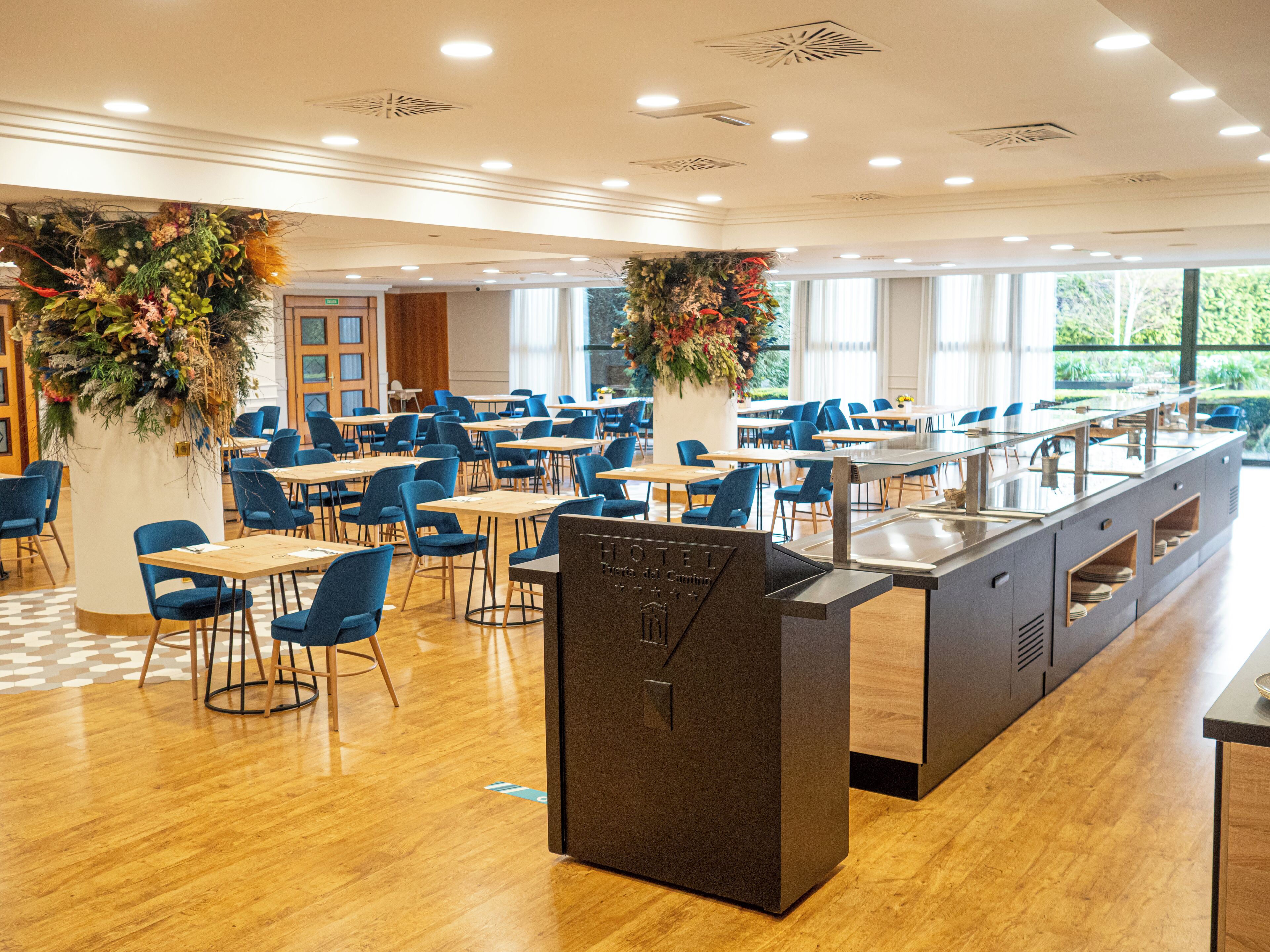 Uma sala de jantar de hotel com várias mesas e cadeiras, um buffet central e amplas janelas que proporcionam luz natural.