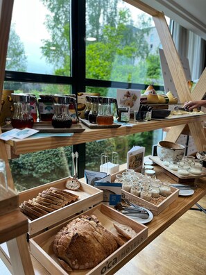 Una barra de madera con diversos alimentos y condimentos, como pan, frascos de miel y queso, con vistas a la vegetación exterior.