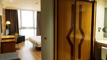 A hotel room with a bed, a desk with a lamp, a wardrobe, and a bathroom with a mirror and sink.