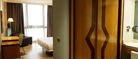 Habitación de hotel con cama, escritorio con lámpara, armario y baño con espejo y lavamanos.