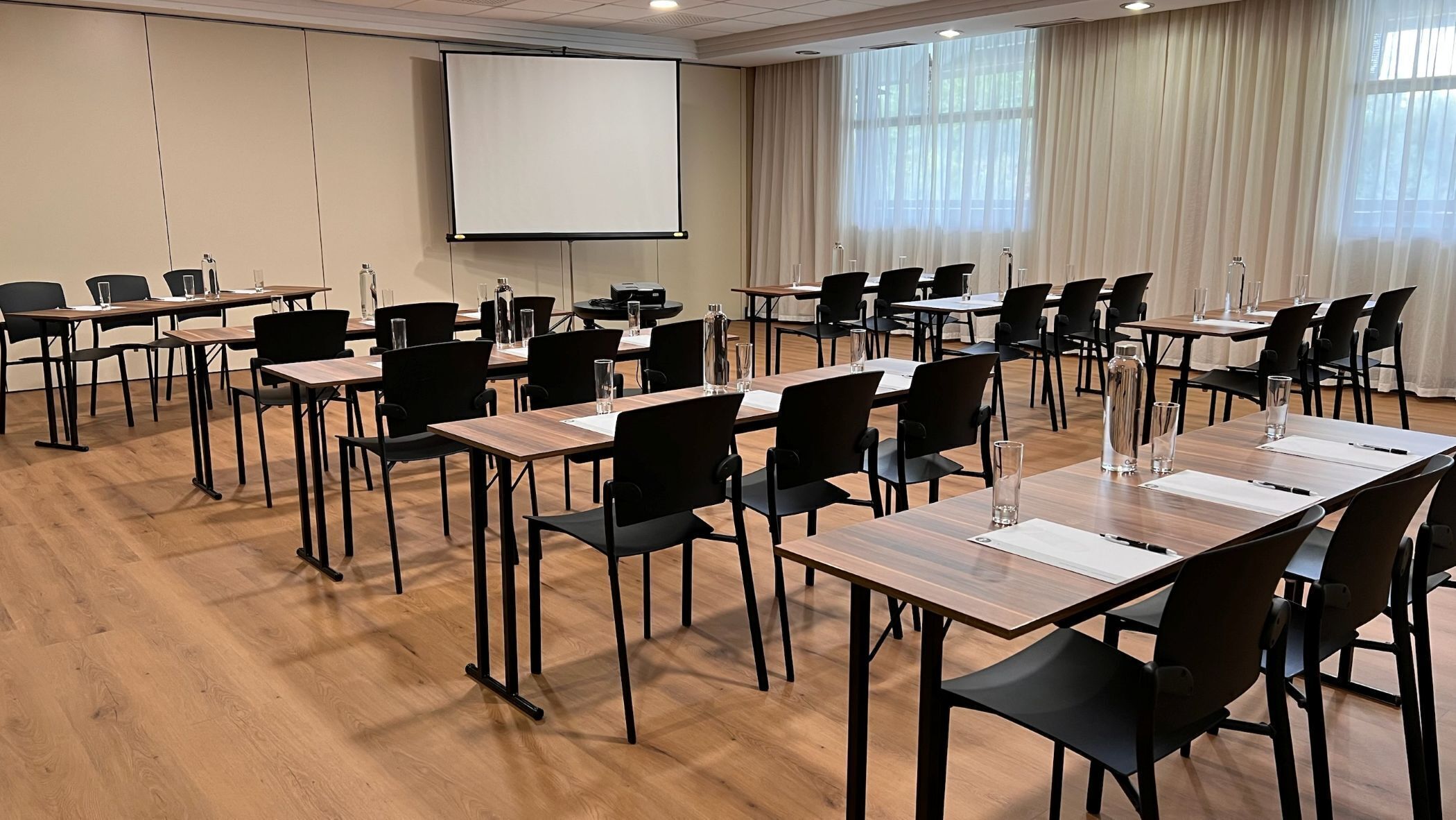 A conference room with multiple tables and chairs, a projector screen, and large windows with curtains.