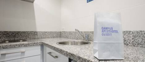 A kitchen with granite countertops, a stainless steel sink, and a white cabinet with drawers. A sign on the wall reads "CAMPUS APARTHOTEL OVIEDO."