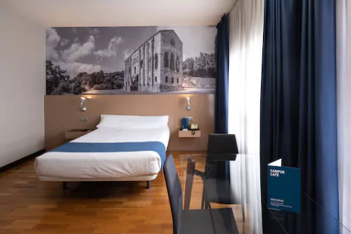 A hotel room with a bed, a desk, a chair, a nightstand, and a wall with a mural.