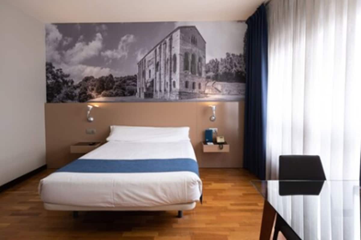 A hotel room with a bed, a nightstand, a wall-mounted lamp, and a window with curtains.