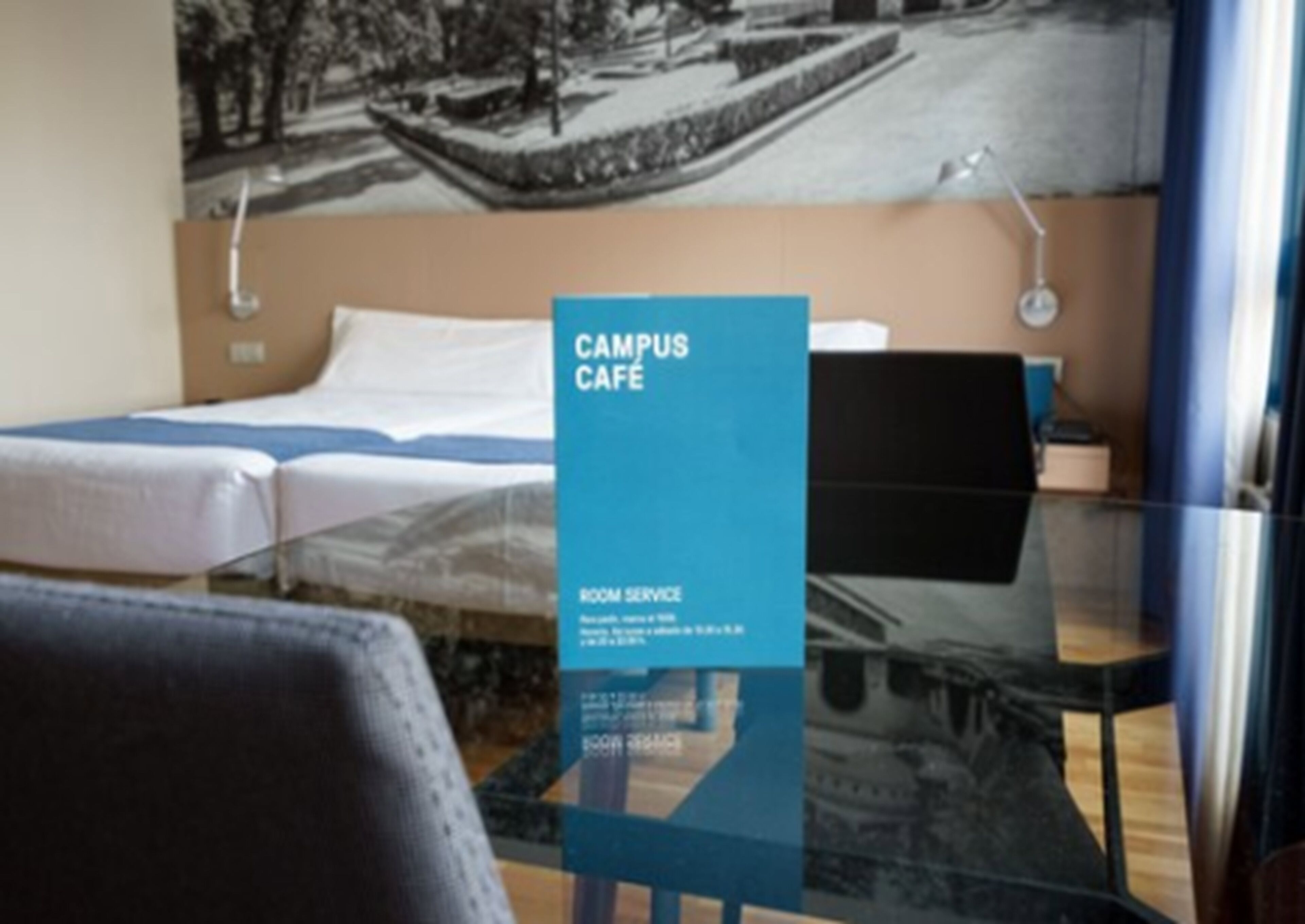 A room with a bed, a desk, and a chair. There is a blue sign that reads "CAMPUS CAFE" and "ROOM SERVICE".
