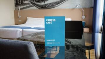 A room with a bed, a desk, and a chair. There is a blue sign that reads "CAMPUS CAFE" and "ROOM SERVICE".