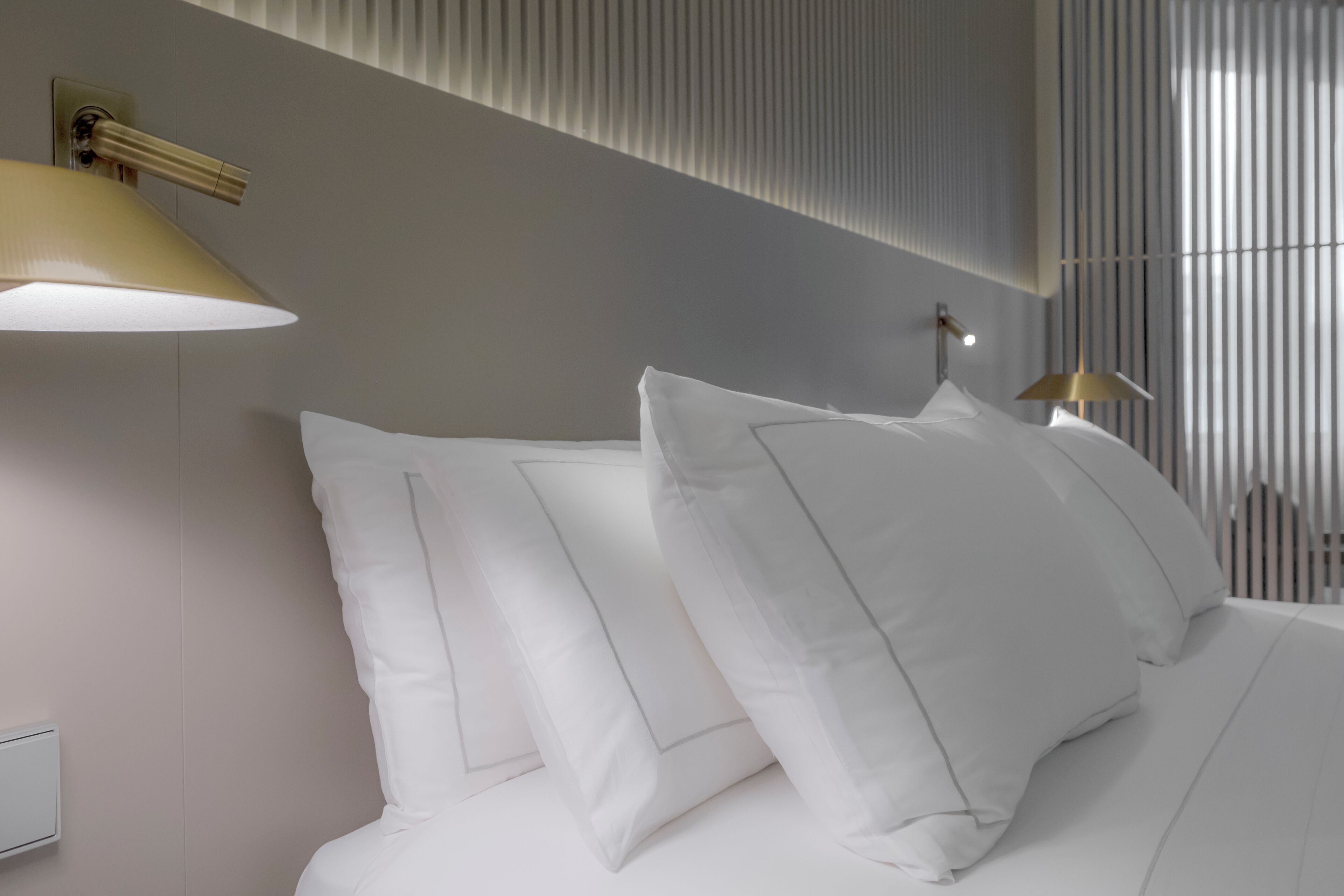 A neatly made bed with white linens and pillows, a wall-mounted light fixture, and a window with vertical blinds.