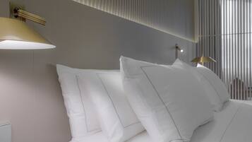 A neatly made bed with white linens and pillows, a wall-mounted light fixture, and a window with vertical blinds.