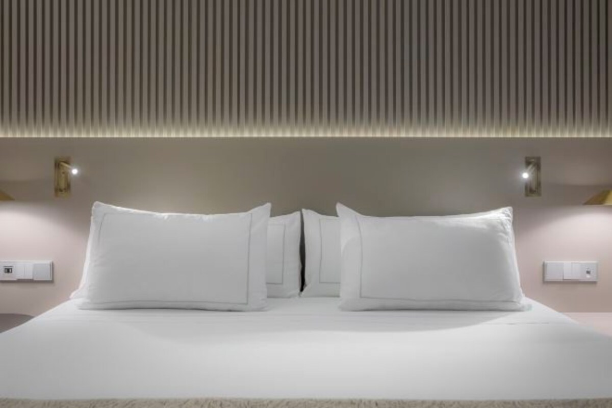 A modern hotel room with a large bed, white bedding, and a wall-mounted light fixture.