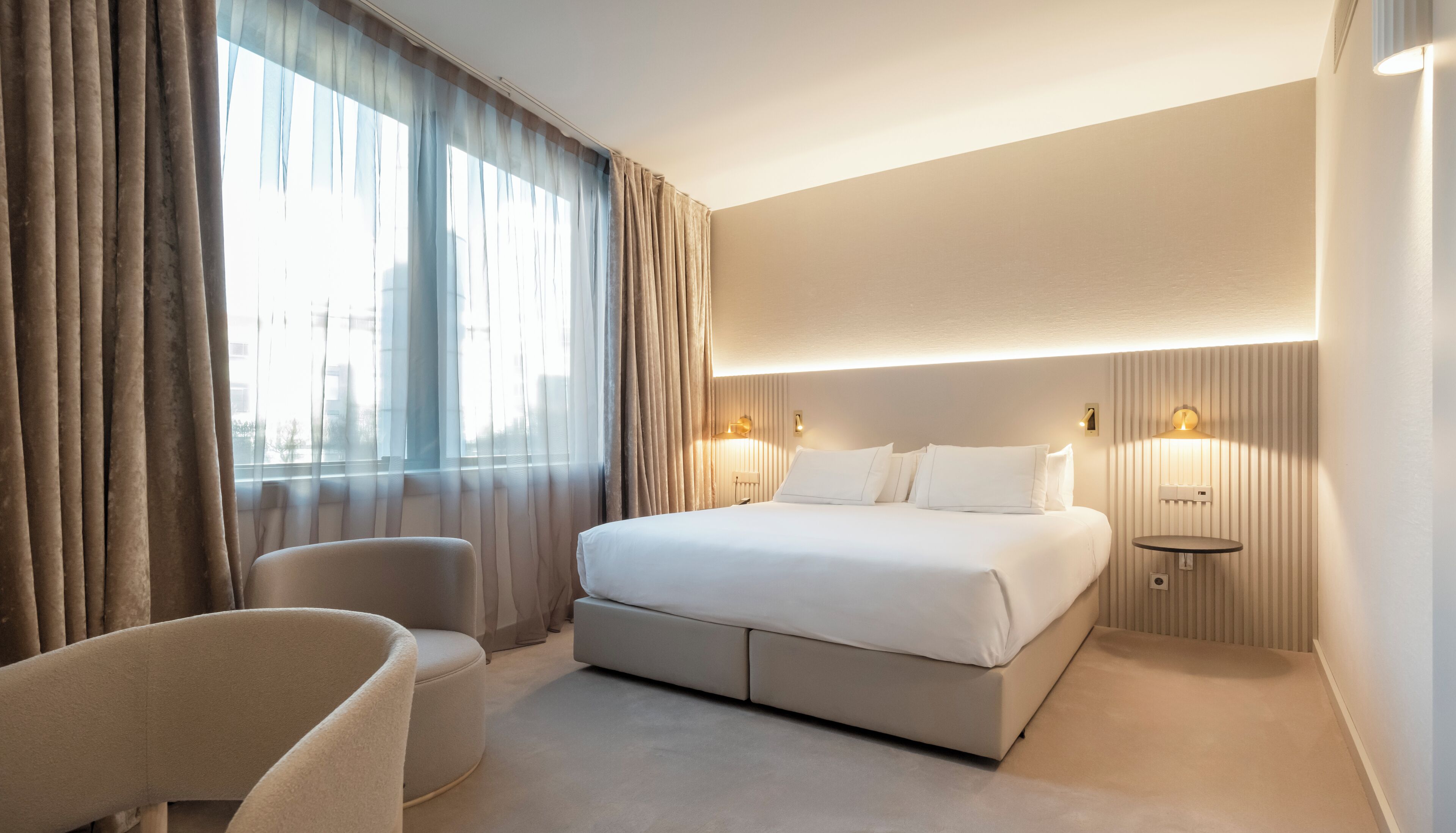 Une chambre d’hôtel moderne avec un grand lit, deux tables de chevet avec des lampes, une chaise et une grande fenêtre avec des rideaux.