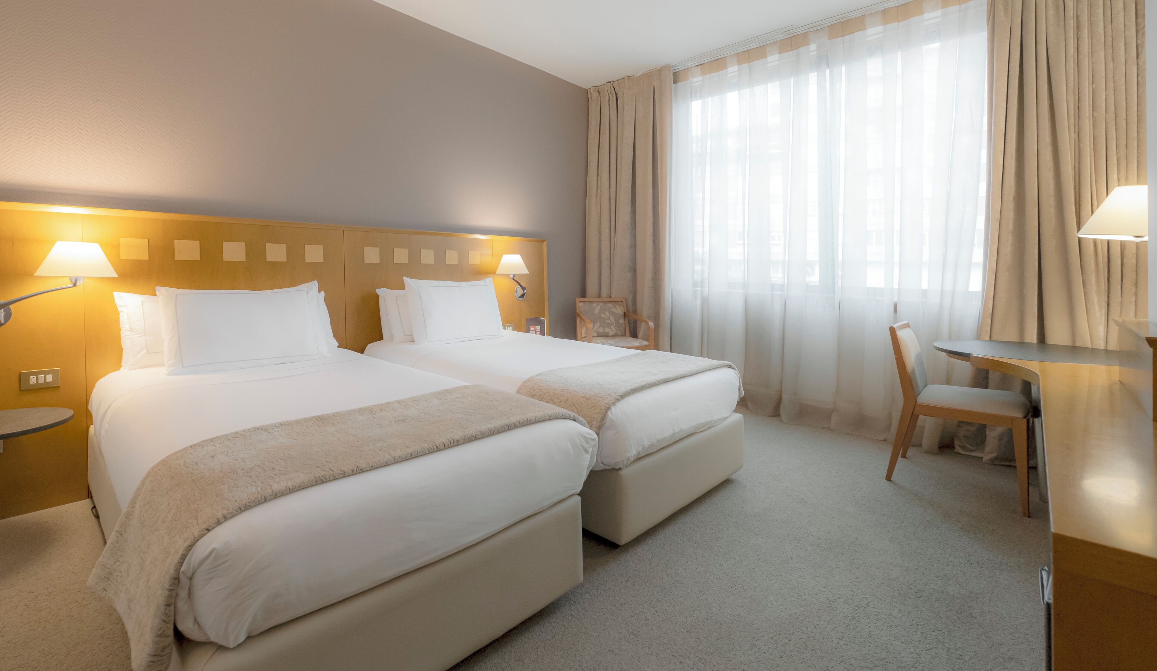 Habitación de hotel con dos camas, un escritorio, una silla y un ventanal con cortinas.
