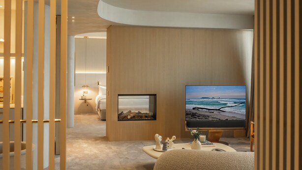 Una habitación de hotel moderna con un televisor de pantalla plana grande mostrando una escena de playa, un rincón de descanso con una mesita pequeña y una cama con una mesita de noche.