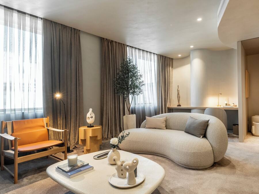 A modern living room with a sofa, a chair, a coffee table with books and a candle, and a large window with curtains.