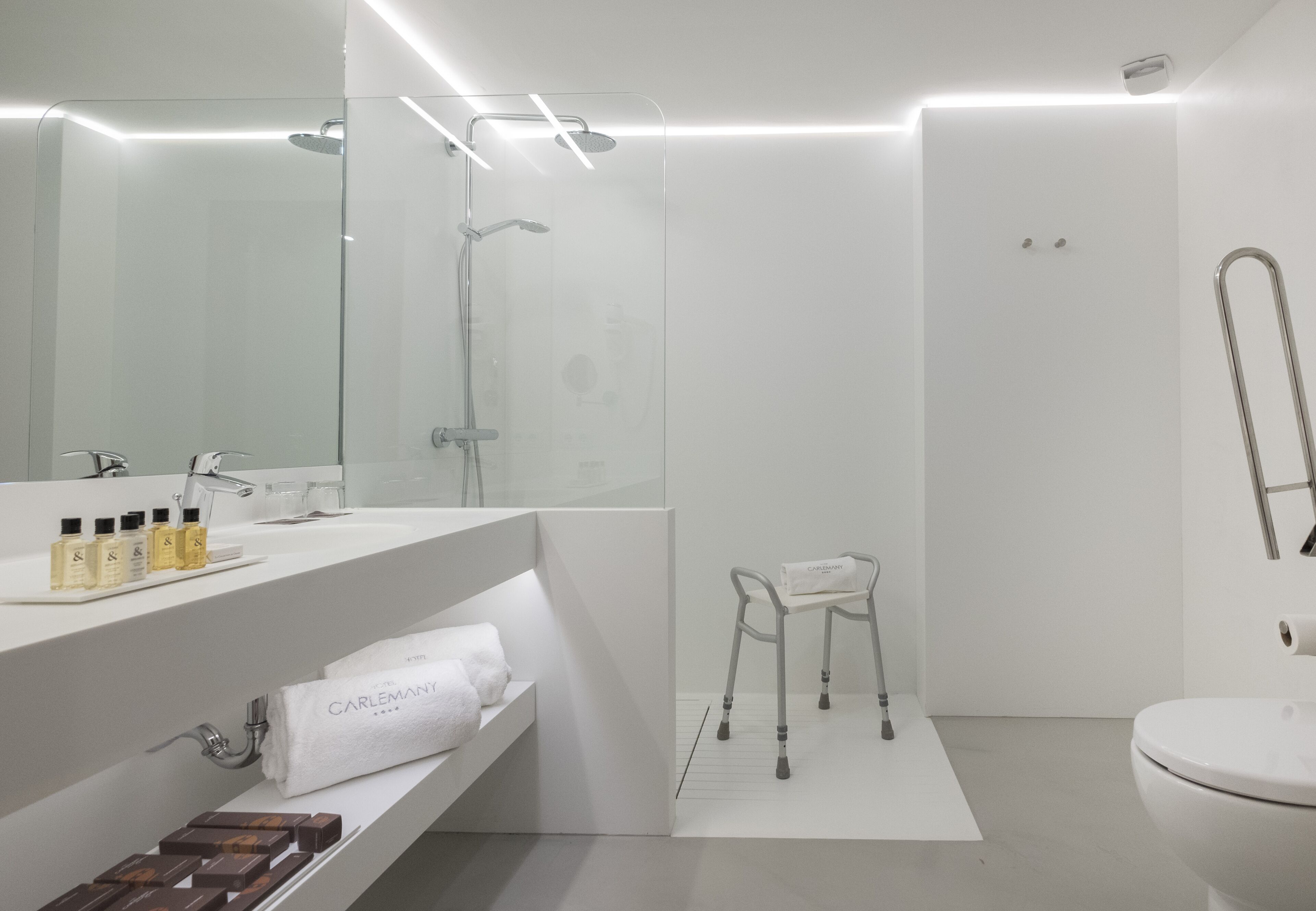 Un baño moderno con ducha envuelta en vidrio, lavamanos blanco con artículos de aseo y una toalla blanca en un estante.