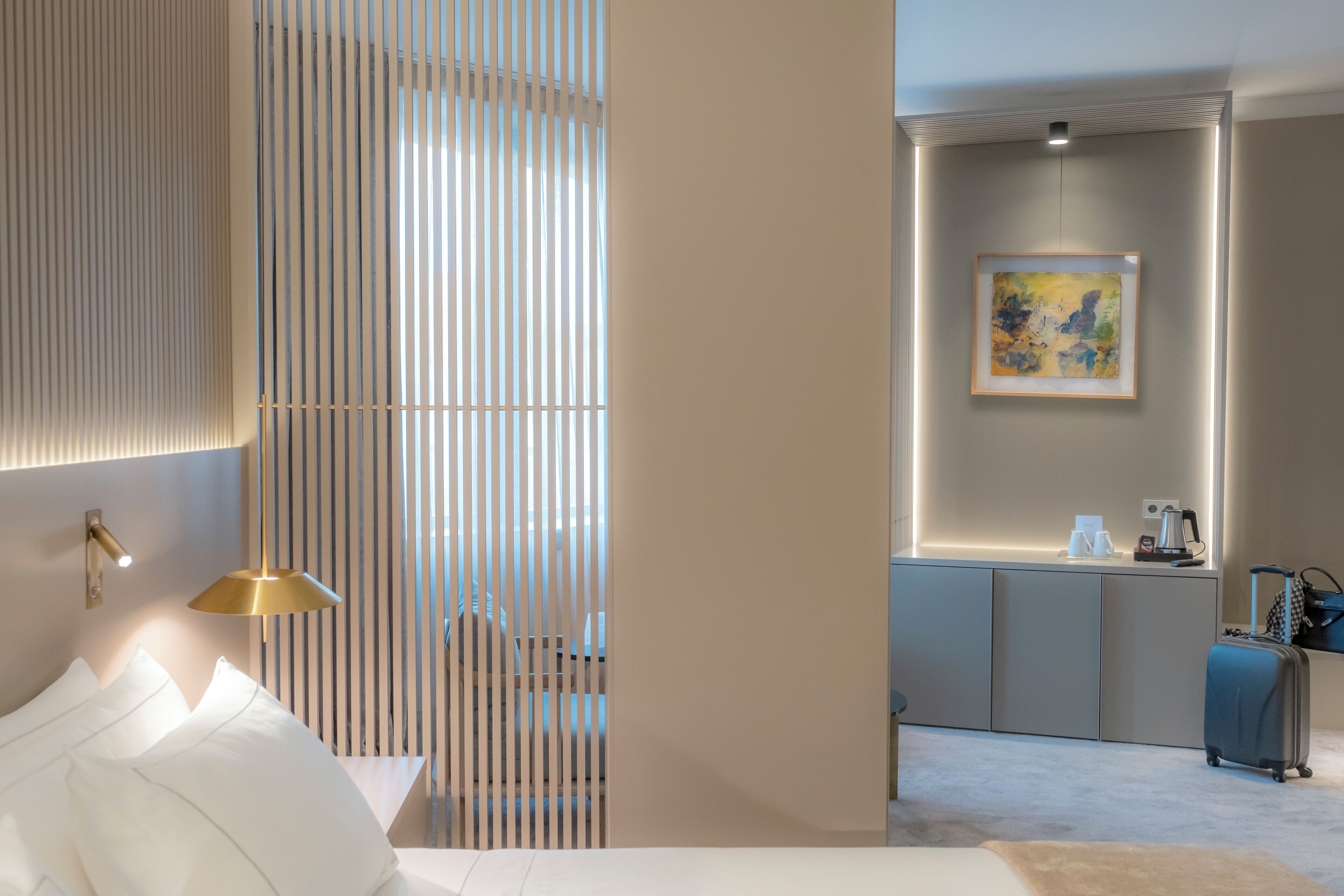 A modern hotel room with a bed, a desk with a lamp, a painting on the wall, and a suitcase.