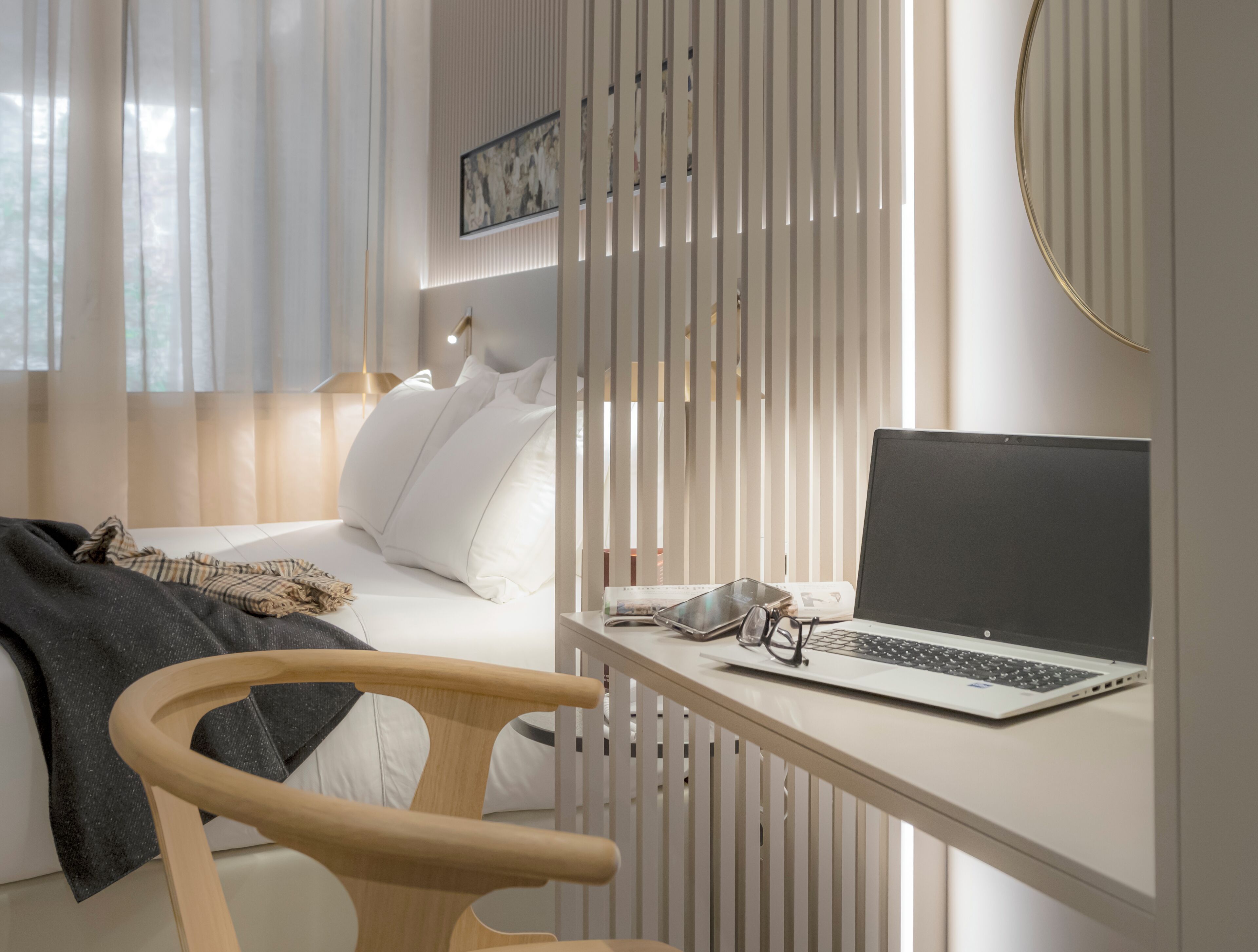 Ein modernes Schlafzimmer mit einem Bett, einem Schreibtisch mit Laptop, einem Stuhl und einem Fenster mit Jalousien.