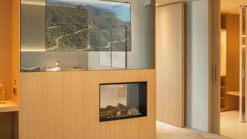 A modern hotel room with a large bed, a wooden console table, a fireplace, and a large window with a mountain view.