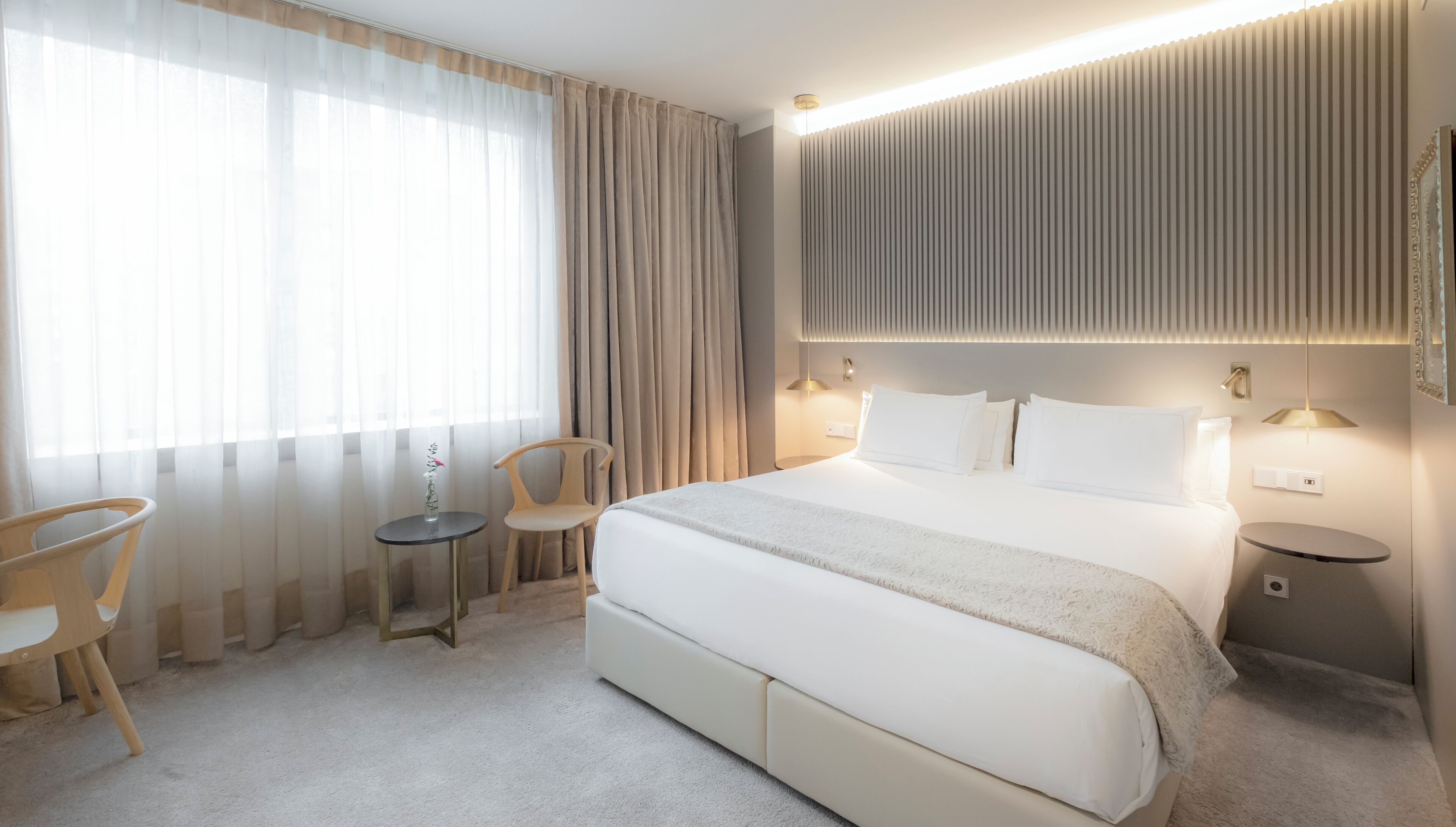 Une chambre d’hôtel moderne, dotée d’un grand lit, de tables de chevet, d’une chaise, d’une petite table et d’une fenêtre avec des rideaux.