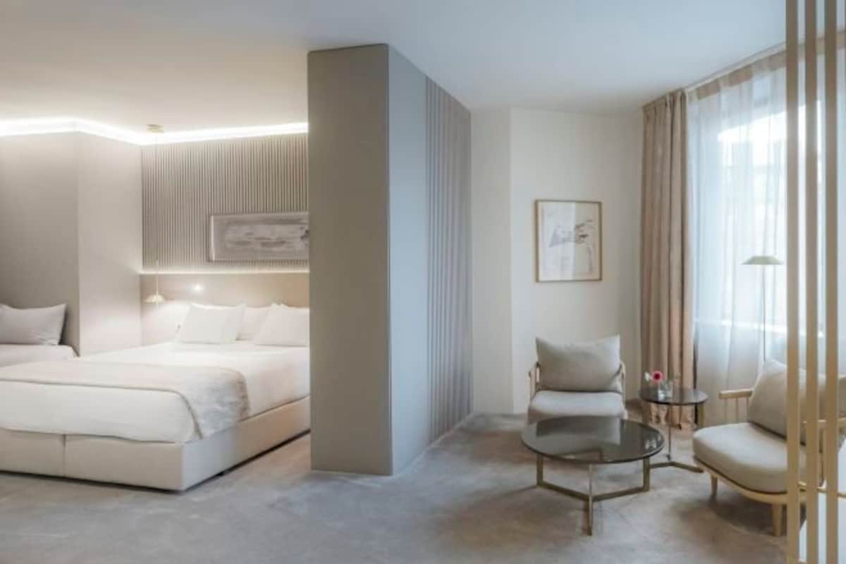 A modern hotel room with a large bed, a seating area with a round table, and a large window with curtains.
