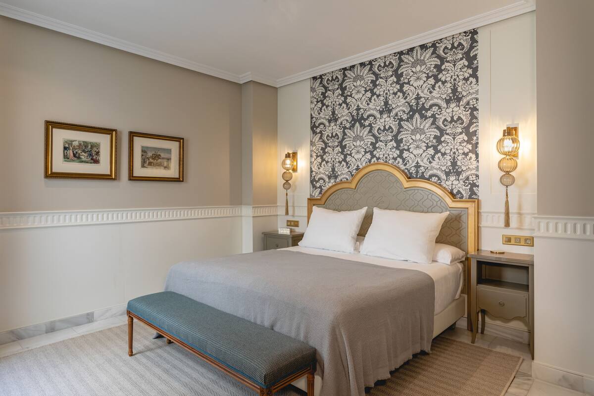 A neatly arranged bedroom with a bed, bench, framed pictures, and wall sconces.