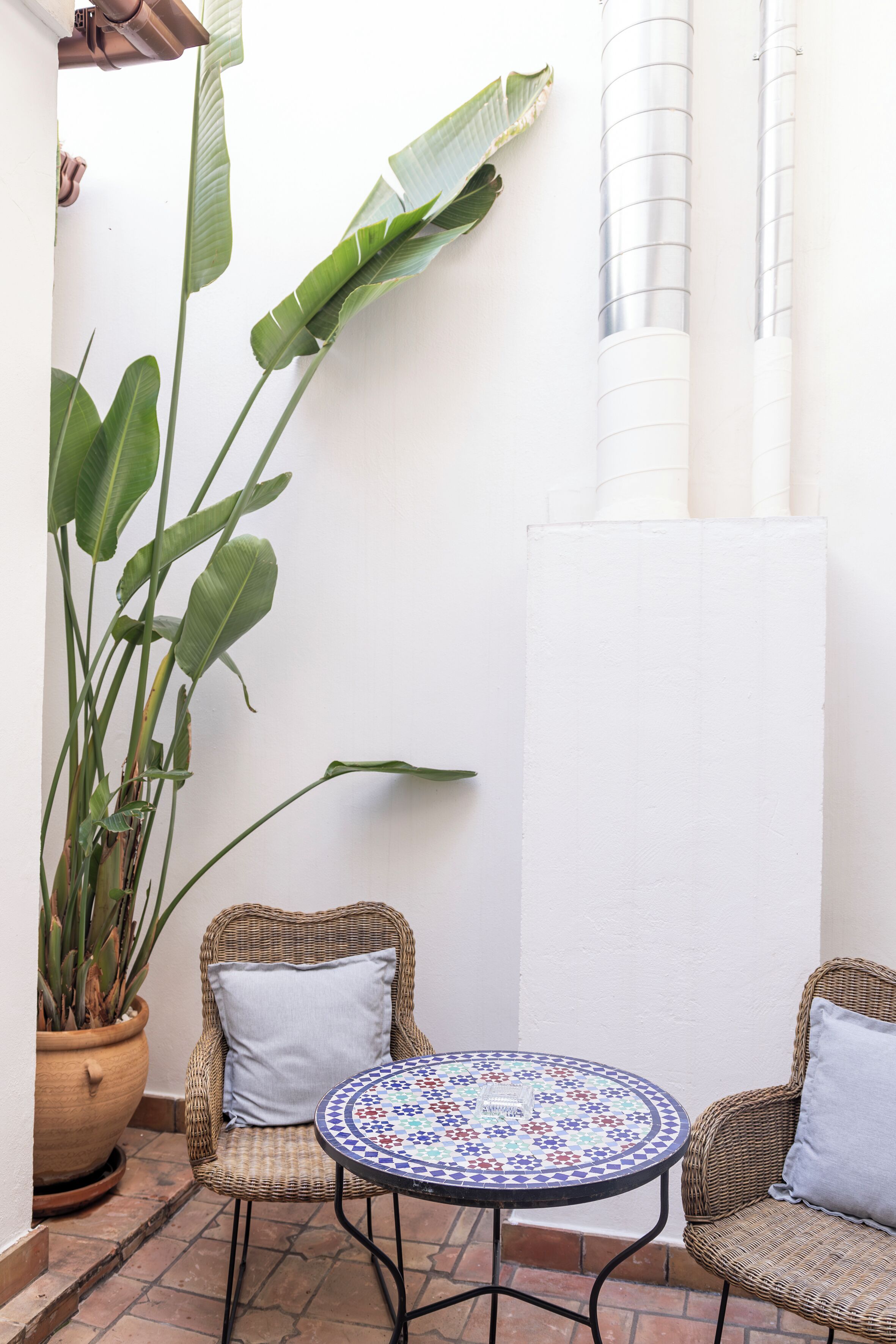 A small patio area with wicker chairs, a round table with a patterned tile top, and a large potted plant.