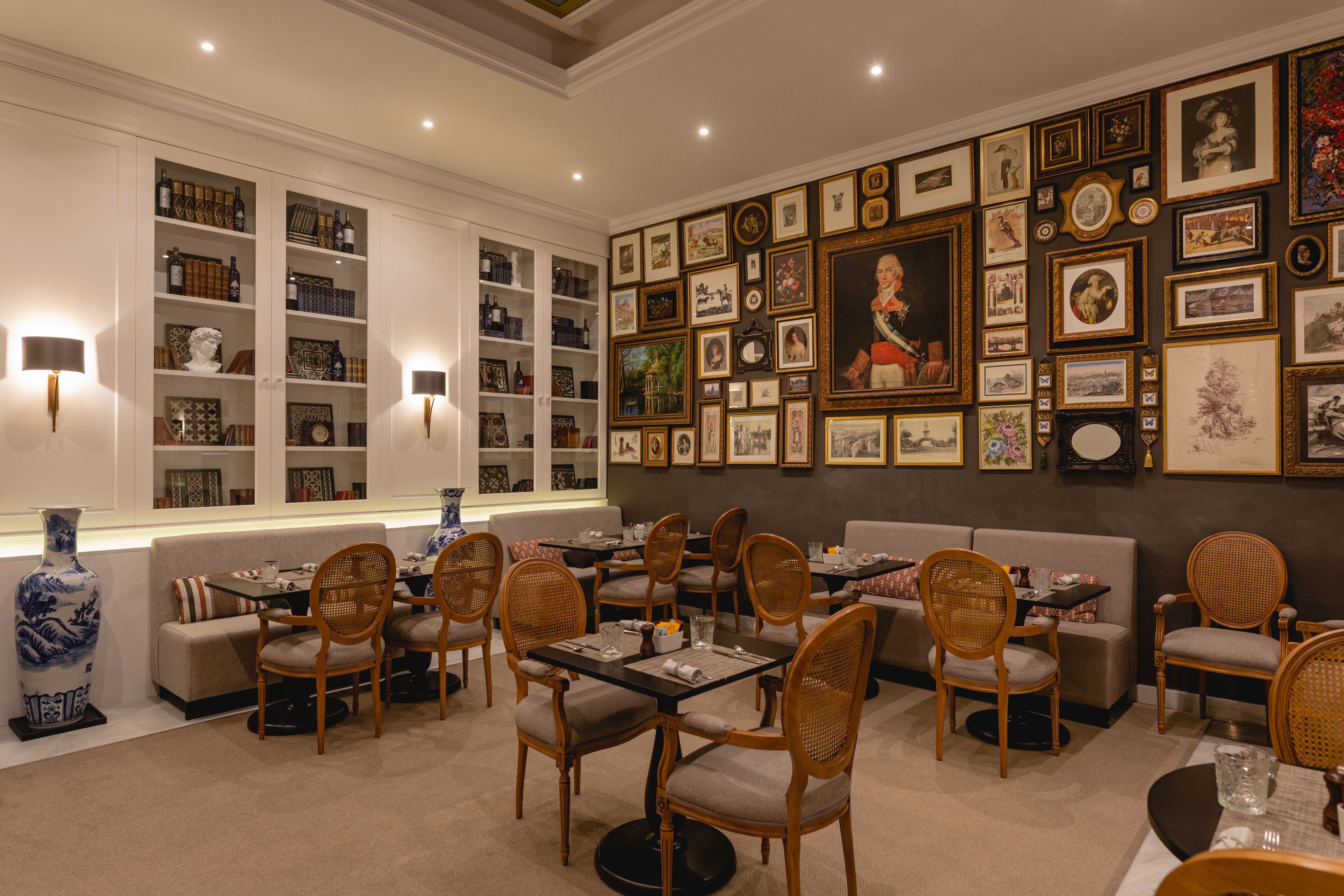 Un espace repas comprenant plusieurs tables et chaises, un grand vase et un mur décoré de tableaux encadrés.