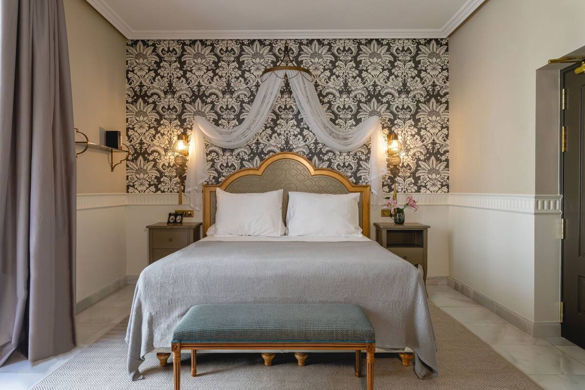 A bedroom with a large bed, two bedside tables, a bench, a wall with a floral pattern, and a door.