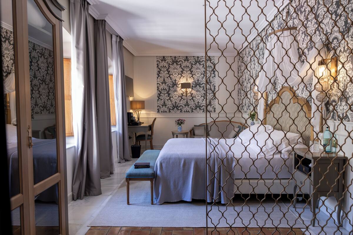 A hotel room with a large bed, a desk, a chair, and a small table with a lamp.